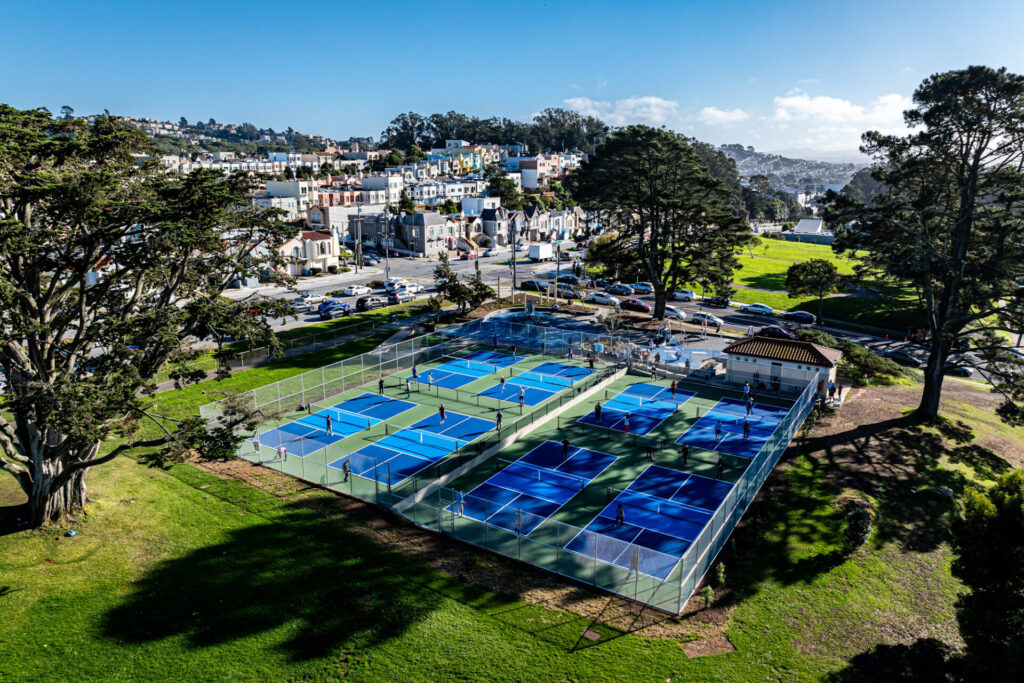 Aerial view of a pickleball court at Carl Larsen park in San Francisco. The pickleball courts were converted from a tennis and basketball court and have been resurfaced using Plexipave acrylic resurfacer in dark blue in the play area, light blue in the kitchen, and medium green on the perimeter with white lines. The courts are surrounded by grass and there are people playing on the courts.