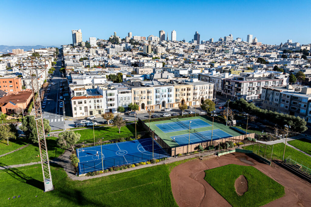 Aerial view of the basketball court, tennis court and multi-use sport court at Moscone Park in San Francisco. The San Francisco city landscape is pictured in the horizon. The courts were refurbished using the slipsheet overlay system and surfaced using Plexipave acrylic resurfacer and color in medium green and dark blue