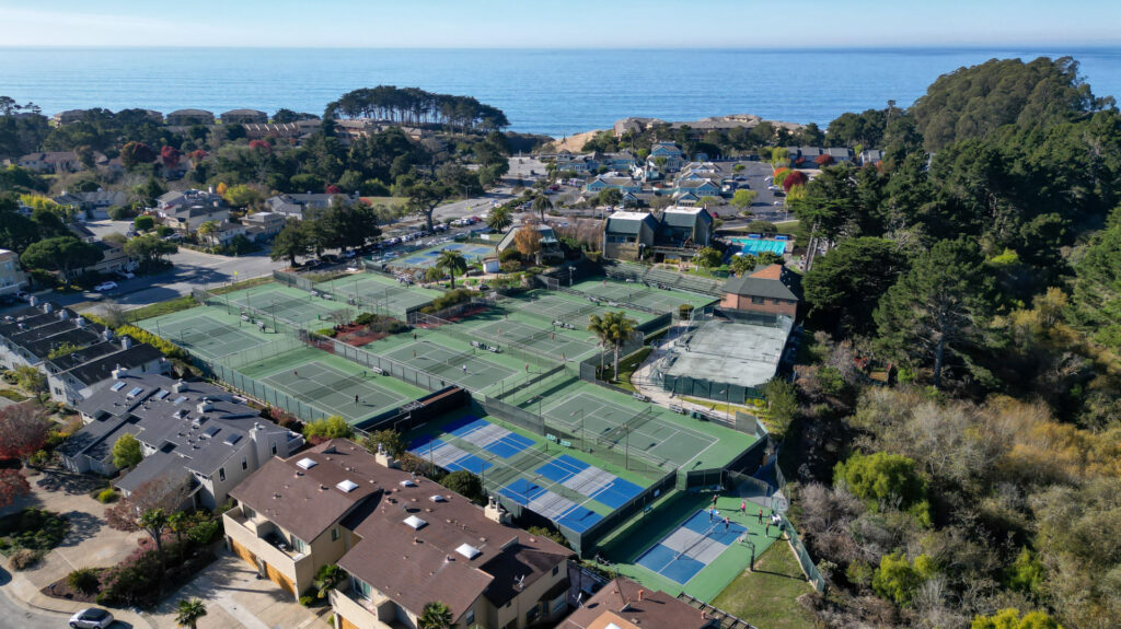 Aerial view of multiple tennis courts at Seascape Sports Club in Aptos, California with the ocean in the background. . Nine of the tennis courts have been resurfaced with Plexipave hardcourt acrylic resurfacer and color in Dark Green and light green. One tennis court has a layout of four pickleball courts painted on top. There is one clay tennis court off to the side and one solo pickleball court towards the bottom.